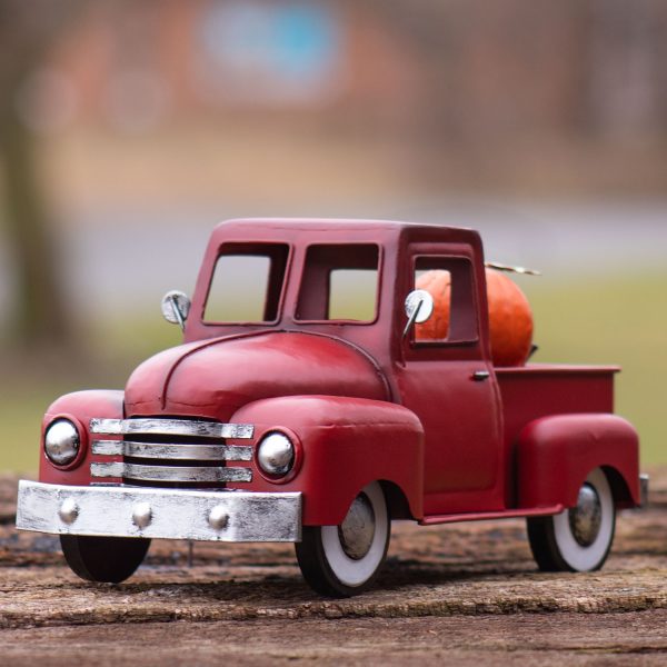 It’s painted in an antique red—deep, slightly weathered, and warm in tone. The body features smooth, rounded edges with a curved front grille and fenders. The truck bed in the back is open and flat, designed to hold small seasonal decorations. The wheels are black with silver centers, and the windows are cutouts with no glass. It has a charming, vintage look with a rustic finish perfect for seasonal displays.