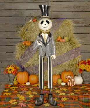 This is a tall Halloween skeleton decoration. He has a big smiling skull, wears a shiny black top hat, a gray jacket with a gold bow tie, and striped pants. He’s holding a stick with a spider web on it. He is standing in front of hay and pumpkins.