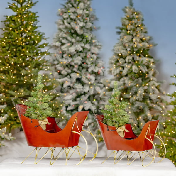 pair of vintage style Christmas sleighs in antique red finish with Christmas trees in it standing in front of lighted up Christmas trees
