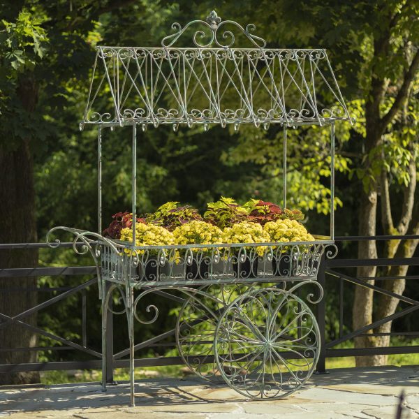 Large iron flower cart with 2 moving wheels holding bright yellow and orange flowers in antique style painted in distressed verdi green finish