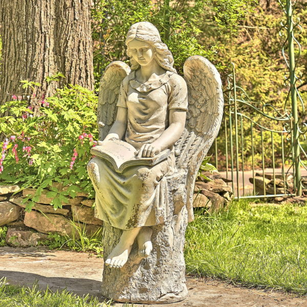 Side view image of Grey angel magnesium statue sitting on a tree stump in the garden reading a book with her head faced down and her wings widely opened behind her