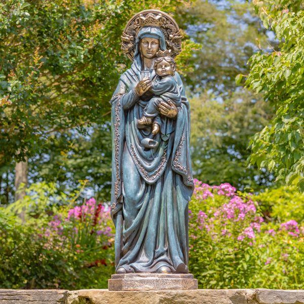 The statue shows a woman standing and gently holding a baby in her arms. She wears long, flowing robes and a head covering. There is a large, round decorative halo behind her head. The baby rests calmly against her chest. The statue looks peaceful, caring, and protective.