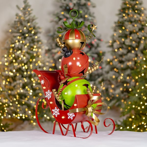 The item is a small decorative red sleigh filled with three large, shiny jingle bells stacked on top of each other like a snowman. The bottom bell is green, the middle one is red with stars, and the top one is gold and red. The sleigh has white snowflakes painted on it and a gold ribbon on the front.