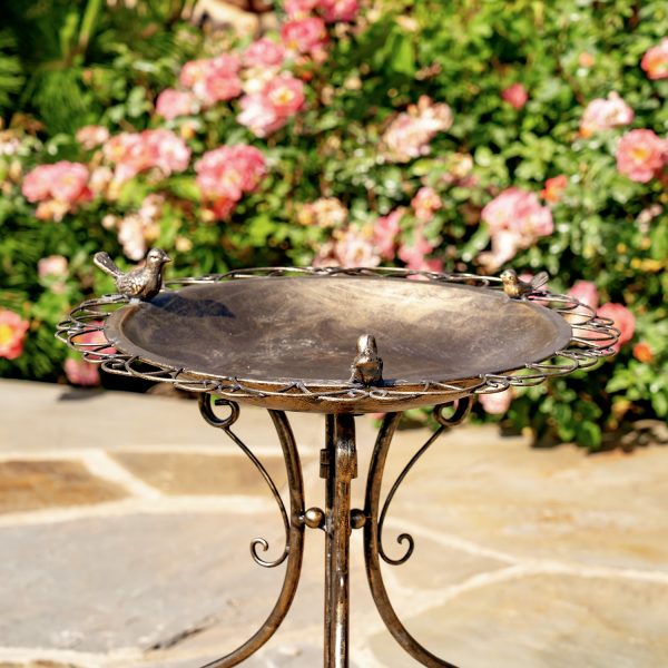 closeup of the basin with 3 small bird sculptures perched on the perimeter in a distressed antique copper-bronze finish - part of a metal birdbath with folding tristand standing on a rock floor surrounded by pink flowers
