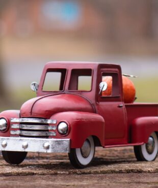 It’s painted in an antique red—deep, slightly weathered, and warm in tone. The body features smooth, rounded edges with a curved front grille and fenders. The truck bed in the back is open and flat, designed to hold small seasonal decorations. The wheels are black with silver centers, and the windows are cutouts with no glass. It has a charming, vintage look with a rustic finish perfect for seasonal displays.
