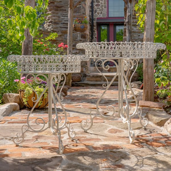 This image shows two white metal plant stands with curly designs standing on a stone path in a garden. There are colorful flowers and green plants all around them.