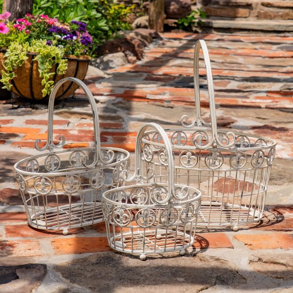 The image shows three white metal baskets of different sizes placed on a stone and brick path. Each basket has a tall, curved handle and decorative swirls along the top edge. They are standing outdoors in the sunlight, with colorful flowers and greenery in the background.