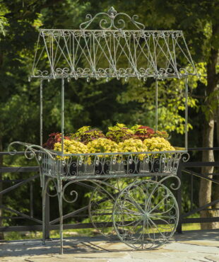 Large iron flower cart with 2 moving wheels holding bright yellow and orange flowers in antique style painted in distressed verdi green finish