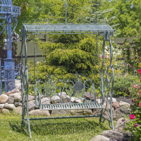 A large iron swing bench in Verdi green with curlicue designs on both ends of the swings , the backrest also has a curlicue design as well as the the top of the sheltered roof on the swing bench