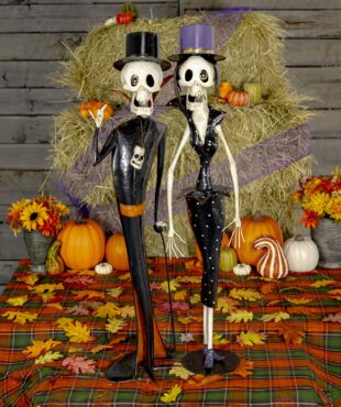 The image shows two tall, thin skeleton figurines dressed in fancy, vintage-style clothing. One is a male skeleton wearing a top hat, black suit with red accents, and holding a cane. The other is a female skeleton in a fitted black dress with white polka dots and a matching top hat. Both have exaggerated features and playful, spooky expressions, like characters from an old horror movie. They are standing side by side against a plain background.