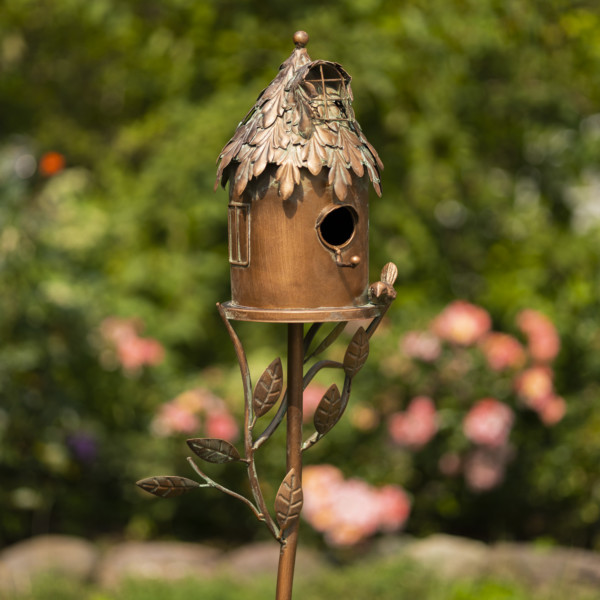 Close up image of Tall bronze to brown country style birdhouse that looks like a round cottage house with bird perched at the rim of the house with a hole for the birds to enter and vines growing from stake coming up to the birdhouse