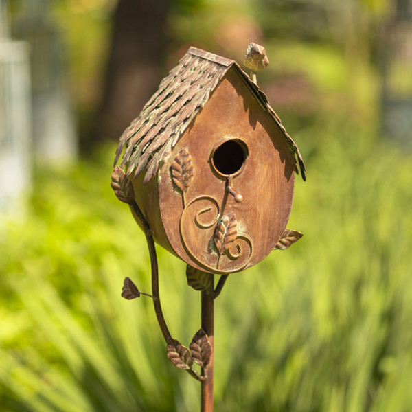 Close up image of Tall bronze to brown country style birdhouse stake that looks like a farm house with small bird perched at the top of the roof with vines growing from stake to the birdhouse where there is a hole for the birds to enter