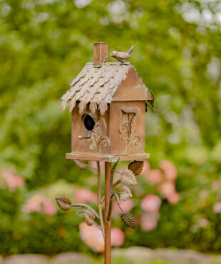 Close up image of the country style bronze to brown birdhouse stake with bird perched at the top of the roof and vines growing from the stake to the house