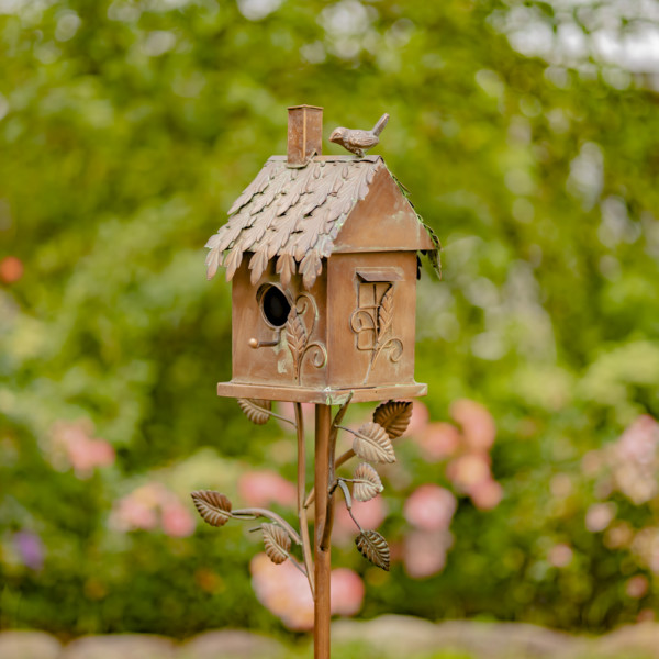 Close up image of the country style bronze to brown birdhouse stake with bird perched at the top of the roof and vines growing from the stake to the house