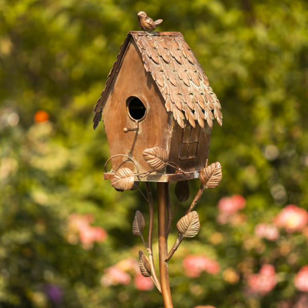 Side close up image of Tall brown to bronze country style birdhouse stake shaped like a cottage house with small bird perched at the top of the roof and vines growing from the stake to the birdhouse and also showing the little hole where the bird can enter