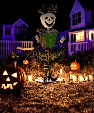 tall iron Halloween skeleton figurine in top hat holding Happy Halloween sign in fall forest surrounded by light up pumpkins and house in purple