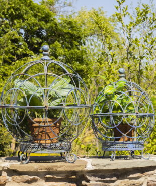 Set of 2 large and small iron globe plant stands in antique blue with plant pots inside on a fence