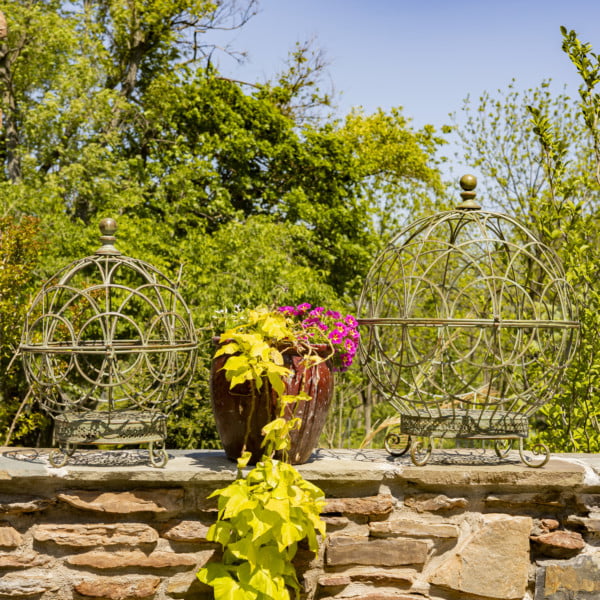 Set of 2 large and small iron globe plant stands in antique green with plant pot between them on a fence