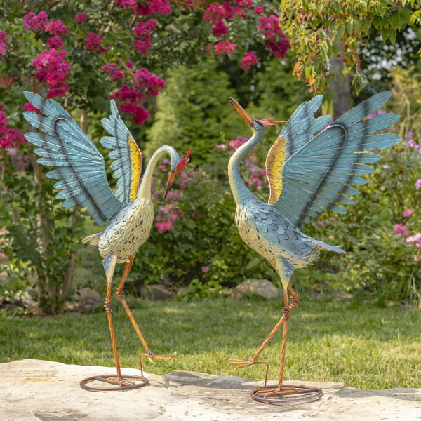 Set of 2 36 inch tall dancing cranes made of iron and hand painted in blue orange and white with distressed finish standing on O shape base in garden