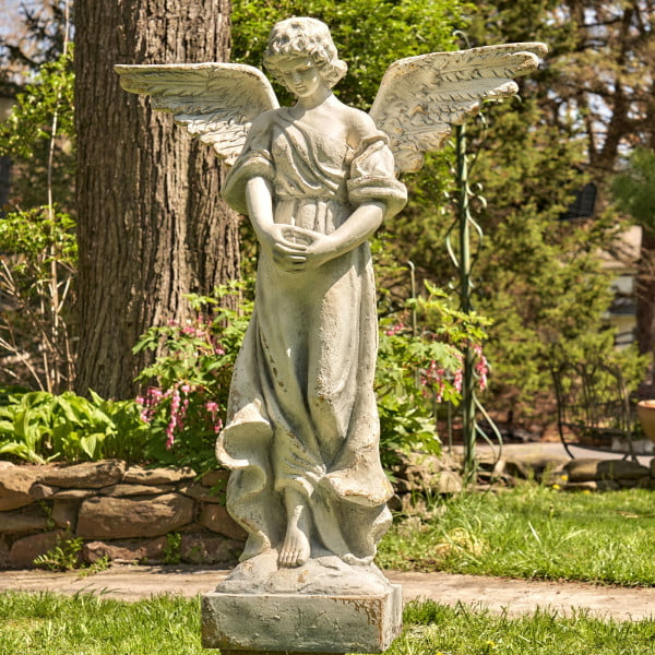 Antique grey tall magnesium angel statue standing in a garden with her head bowed down holding her two hands together while her wings are spread wide open