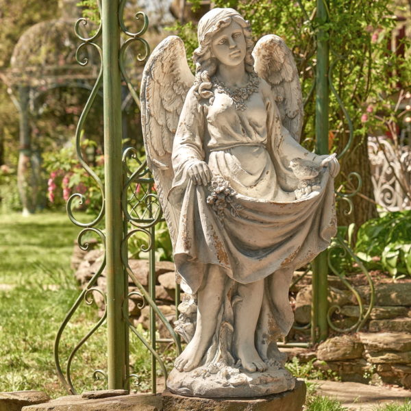 Antique grey tall angel magnesium statue holding up her dress creating a birdbath while a bird is perched at the edge of her dress and she is admiring it with two wings folded behind her