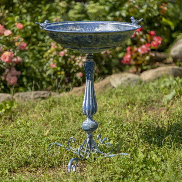 27 inch tall iron birdbath with round basin with two small bird sculptures perched on each side and a decorative pedestal stand and four flourish style legs hand painted in a distressed Antique blue finish outside with pink flowers in background