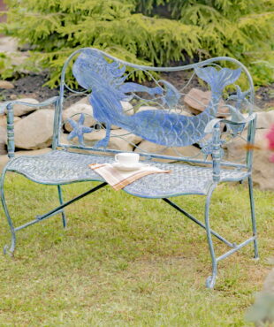 full product image of Sirena Mermaid Coastal Iron Garden Bench in backyard with teacup and plaid cloth napkin resting on bench and hardscaping and pink flowers in background