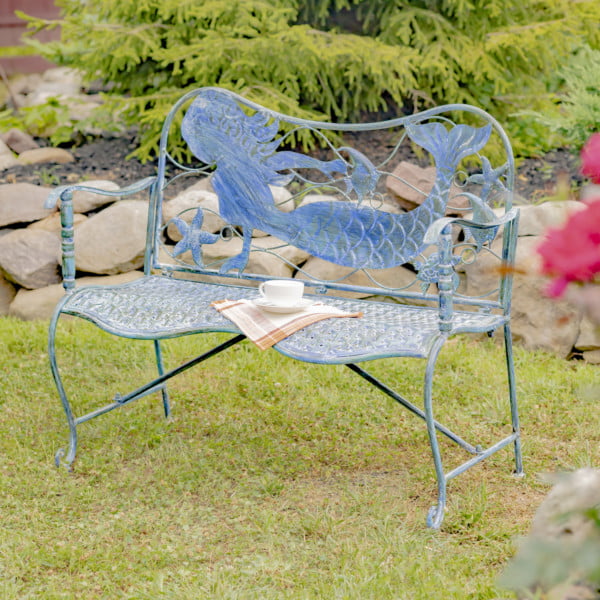 full product image of Sirena Mermaid Coastal Iron Garden Bench in backyard with teacup and plaid cloth napkin resting on bench and hardscaping and pink flowers in background