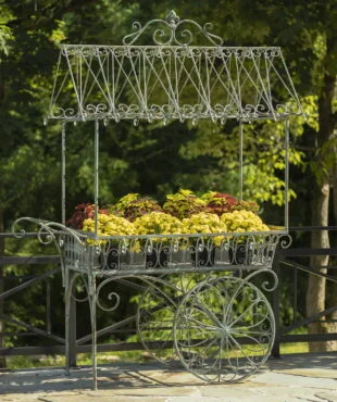 Large iron flower cart with 2 moving wheels holding bright yellow and orange flowers in antique style painted in distressed verdi green finish