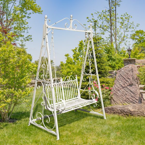 iron swing bench with ornate details in distressed antique white finish standing in beautiful garden