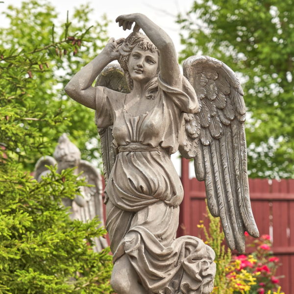 Close up image of Tall grey angel statue standing on a pillar with her two arms up and one feet up balancig on the other one with her long flowing gown and her wings widely opened