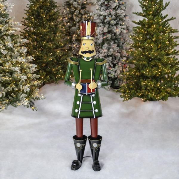 Life-size iron Christmas nutcracker figure Leo in green holding drum in front of lighted up trees