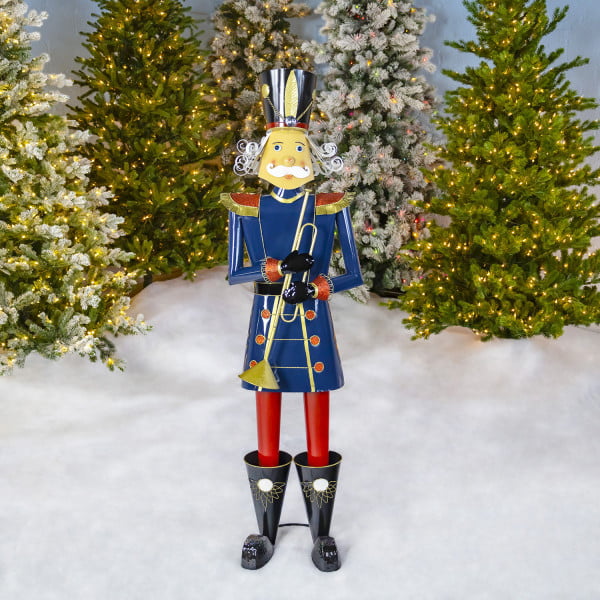 Life-sized iron Christmas blue nutcracker Harold holding trumpet in front of lighted up Christmas trees