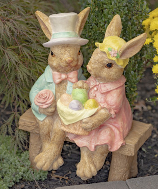 Fancy Rabbit Couple magnesium statue sitting on a bench ,one holding a basket of colorful eggs while the other is in his suite holding a rose