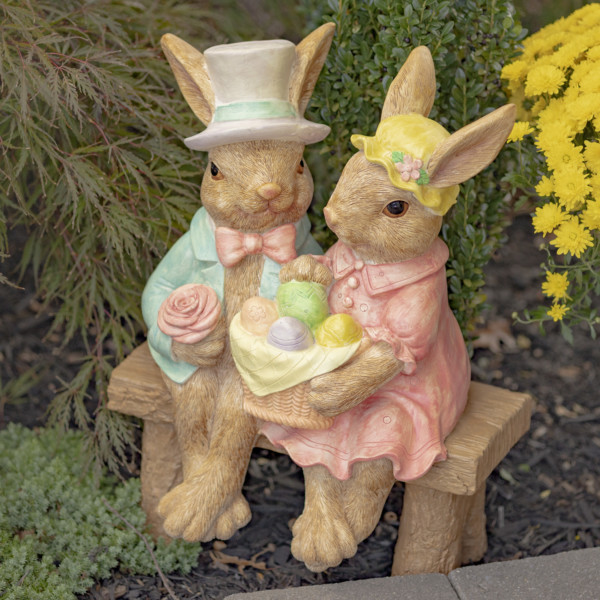 Fancy Rabbit Couple magnesium statue sitting on a bench ,one holding a basket of colorful eggs while the other is in his suite holding a rose