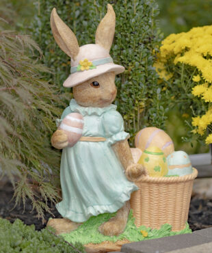 Side view of Lady Easter rabbit garden statue in blue dress and pink sun hat holding an Easter egg in one hand and the other dragging a basket filled with them