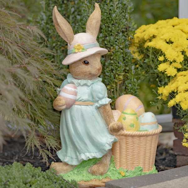 Side view of Lady Easter rabbit garden statue in blue dress and pink sun hat holding an Easter egg in one hand and the other dragging a basket filled with them
