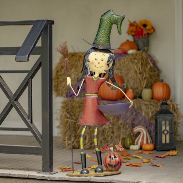 Halloween Decoration of Tall Iron Witch girl in red dress holding a broom in one hand and in other holding a bowl with pumpkin by her feet on the floor