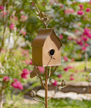 Close up image of Tall antique copper birdhouse stake with a circular hole for birds to go into with vines running from the stake to the birdhouse with a sloped roof