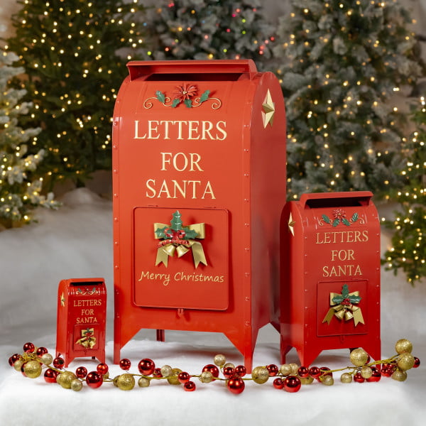 Three Red Santa mailboxes in small medium and large sizes with gold accents and bells in front of light up Christmas trees