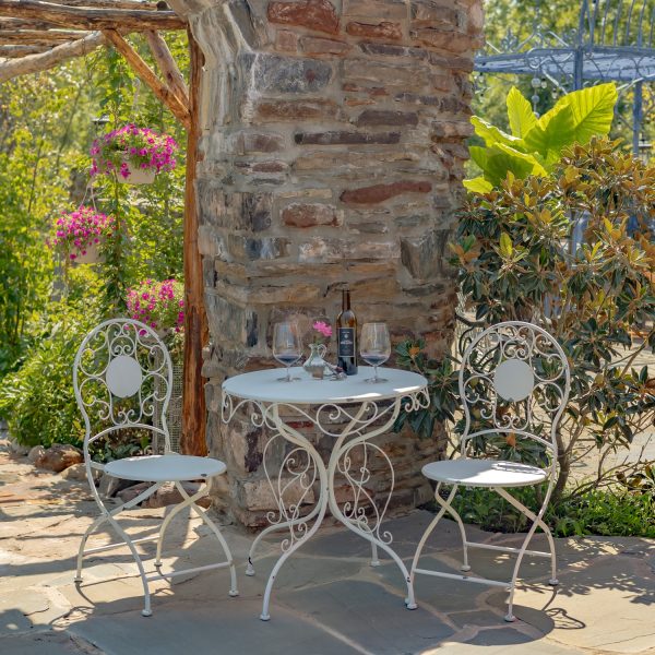 Antique white metal bistro set that includes two chairs and a table with curlicue designs, with wine bottle and 2 glasses standing on the table