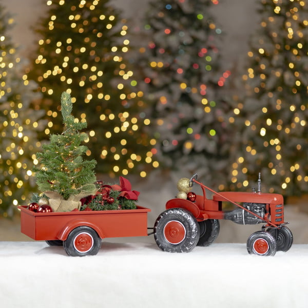 Small red metal tractor with Christmas tree in wagon and large black wheels showing tractor driver seat standing on white snow