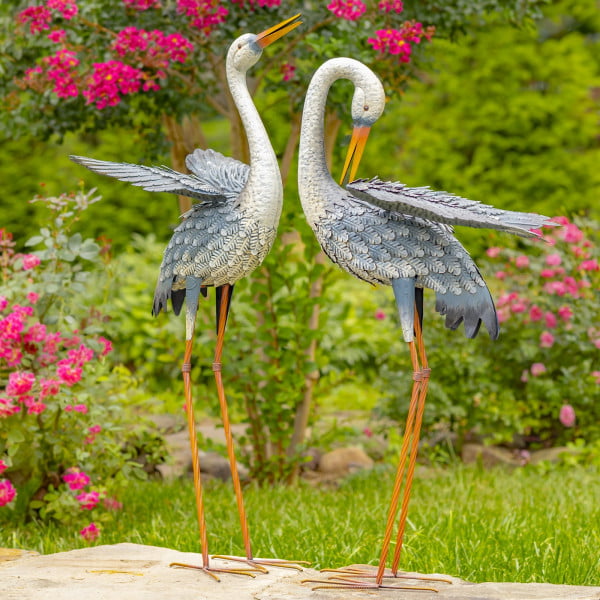 Set of two blue and white egret garden figurines with one looking to the back of him with his neck bent while he is trying to scratch himself while the other one is looking forward standing tall and upright