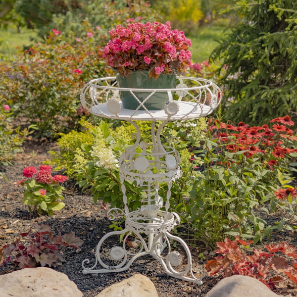 Iron round Victorian style planter table featuring aster blossom and rope-like twisted metal pattern in antique white distressed finish with pink potted flowers in garden