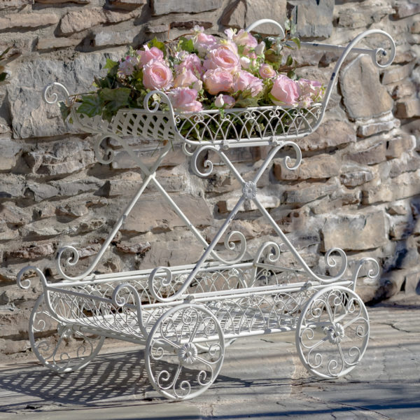 Antique white two tier flower push cart with four moving wheels and curlicue designs with pink flowers in it ( Flowers not included)