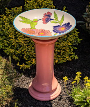 24 inch tall porcelain pedestal birdbath with peach base and painted green hummingbird with large flowers on white basin in garden