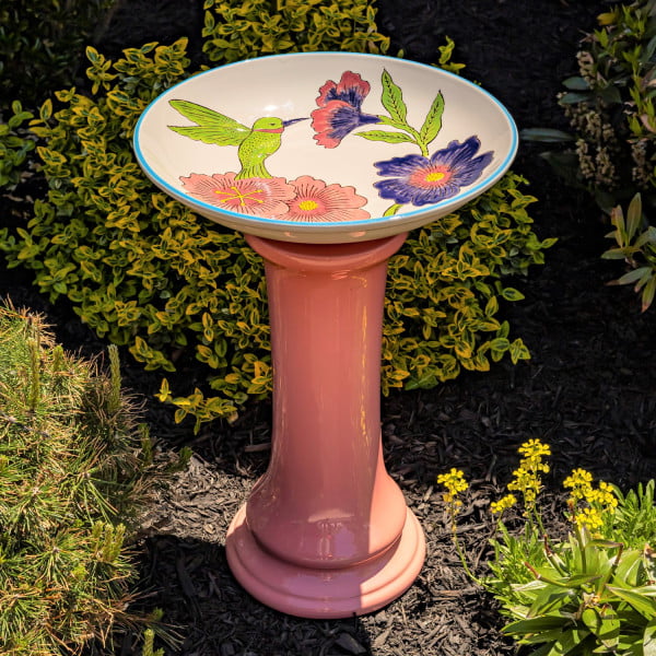 24 inch tall porcelain pedestal birdbath with peach base and painted green hummingbird with large flowers on white basin in garden
