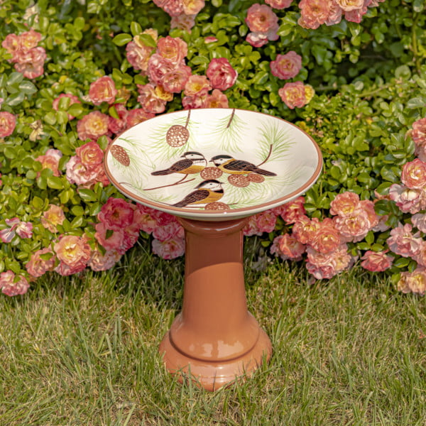 15″ Tall Round Porcelain Pedestal Birdbath with Hand Painted Birds in a Pine Tree “Finch” in front of rose bush