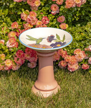 15 inch tall round porcelain pedestal birdbath in peach with hand painted hummingbird and large flower blossoms “Hadley” in front of rose bush