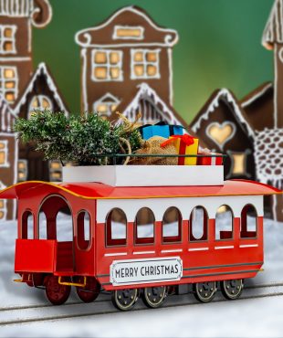 Full Length Image of 20 inch Long Vintage Style Metal Christmas Trolley with Tree & Gifts on Roof Slightly Turned and Displayed on Tracks in Front of a Beautiful Illuminated Gingerbread Village